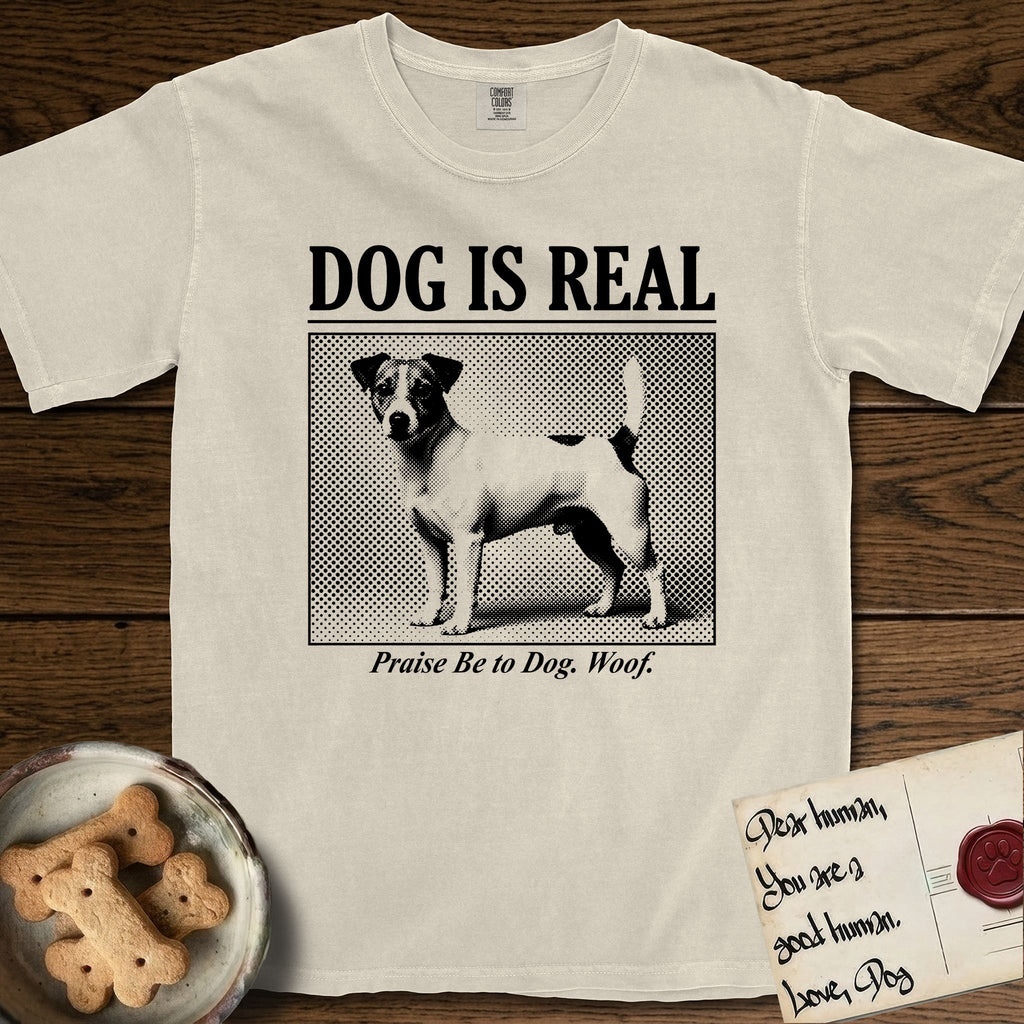 Dog is Real Jack Russell T-Shirt flat lay in Ivory, featuring Black halftone newspaper photo of a mostly white Jack Russell with dark ears and an upright tail standing in profile with head turned to the camera, under the headline DOG IS REAL, with the caption Praise Be to Dog. Woof..