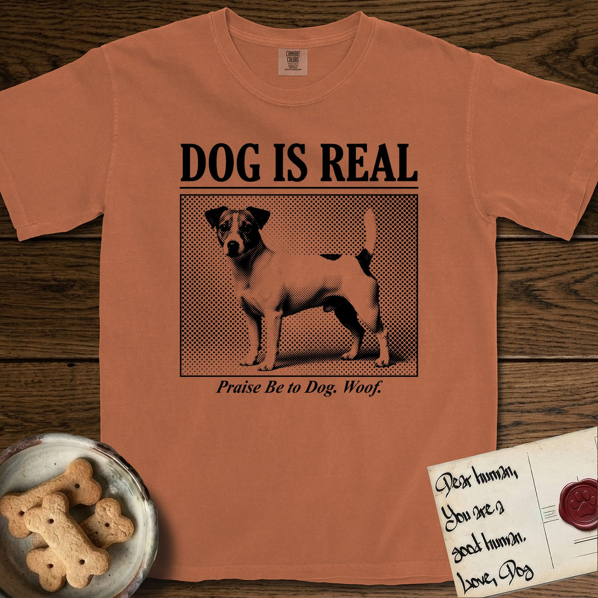 Dog is Real Jack Russell T-Shirt flat lay in Yam, featuring Black halftone newspaper photo of a mostly white Jack Russell with dark ears and an upright tail standing in profile with head turned to the camera, under the headline DOG IS REAL, with the caption Praise Be to Dog. Woof..