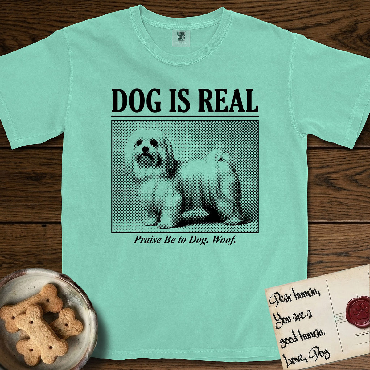 Dog is Real Maltese T-Shirt flat lay in Island Reef, featuring Black halftone newspaper photo of a long-coated white Maltese standing in profile with head turned to the camera, under the headline DOG IS REAL, with the caption Praise Be to Dog. Woof..
