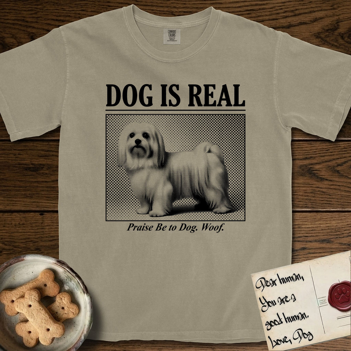Dog is Real Maltese T-Shirt flat lay in Khaki, featuring Black halftone newspaper photo of a long-coated white Maltese standing in profile with head turned to the camera, under the headline DOG IS REAL, with the caption Praise Be to Dog. Woof..