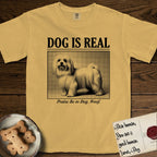 Dog is Real Maltese T-Shirt flat lay in Mustard, featuring Black halftone newspaper photo of a long-coated white Maltese standing in profile with head turned to the camera, under the headline DOG IS REAL, with the caption Praise Be to Dog. Woof..
