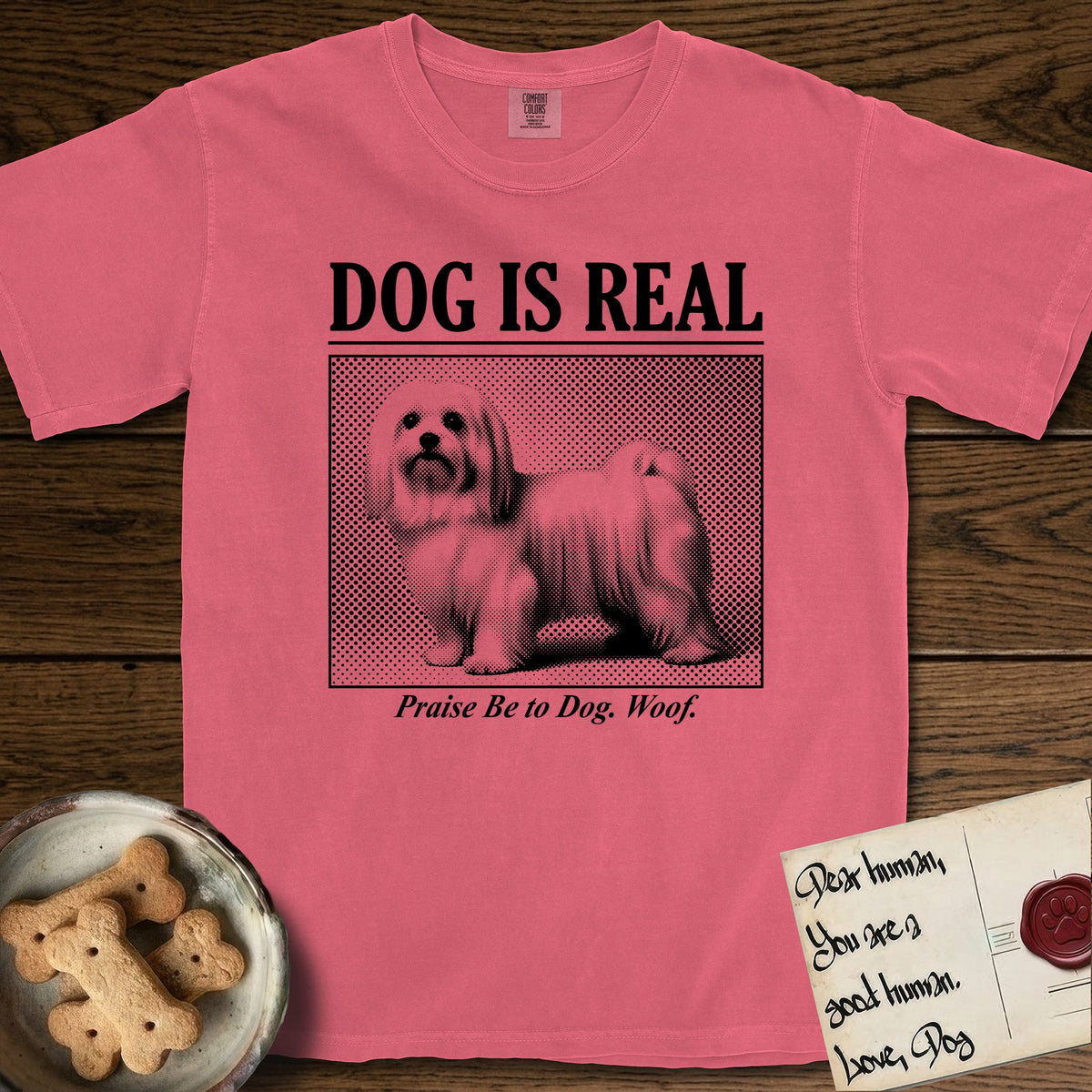 Dog is Real Maltese T-Shirt flat lay in Watermelon, featuring Black halftone newspaper photo of a long-coated white Maltese standing in profile with head turned to the camera, under the headline DOG IS REAL, with the caption Praise Be to Dog. Woof..