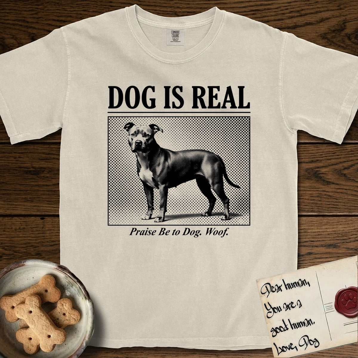 Dog is Real Pitbull T-Shirt flat lay in Ivory, featuring Black halftone newspaper photo of a broad-chested Pitbull standing in profile with head turned to the camera, under the headline DOG IS REAL, with the caption Praise Be to Dog. Woof..