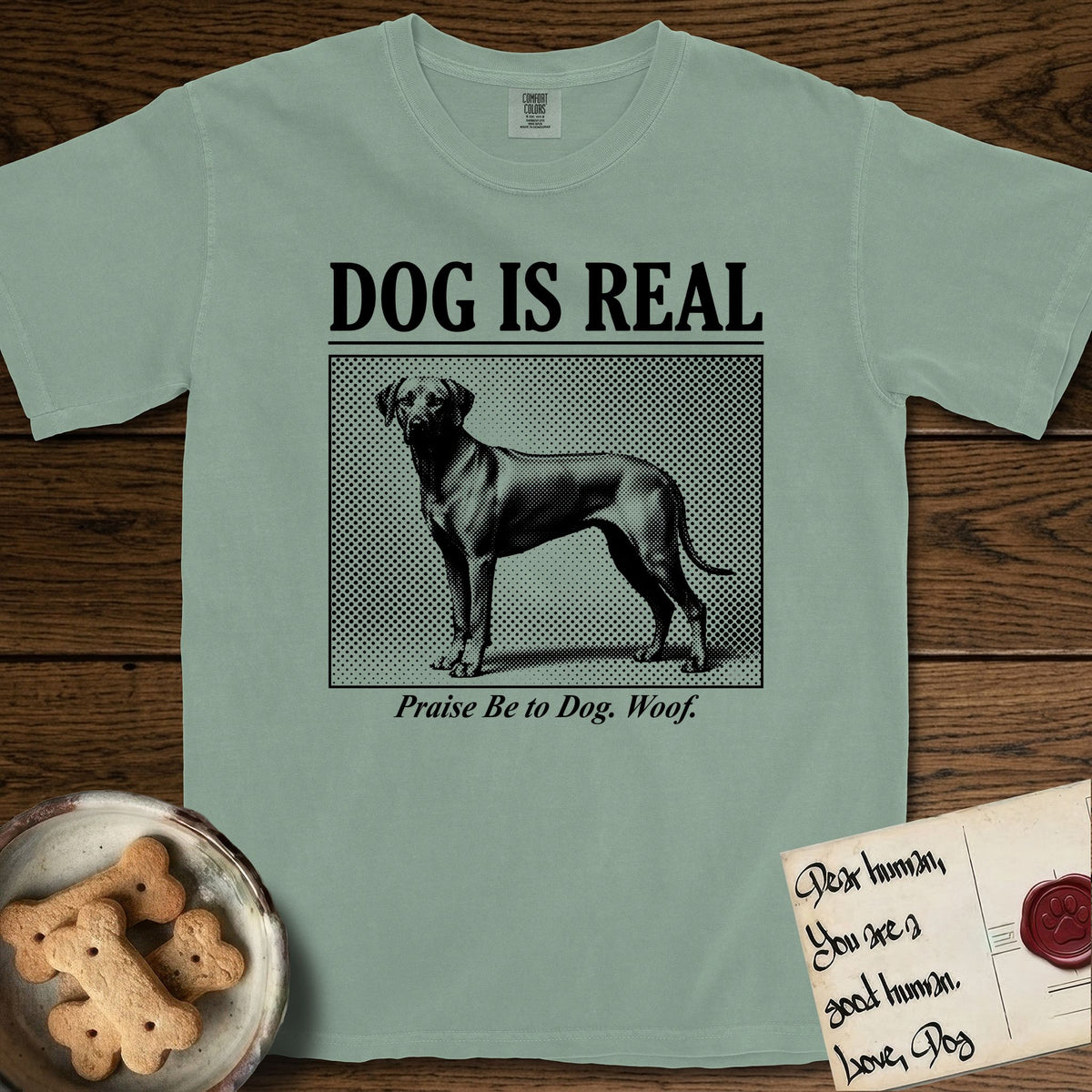 Dog is Real Rhodesian Ridgeback T-Shirt flat lay in Bay, featuring Black halftone newspaper photo of a lean Rhodesian Ridgeback standing in profile with head turned to the camera, under the headline DOG IS REAL, with the caption Praise Be to Dog. Woof..