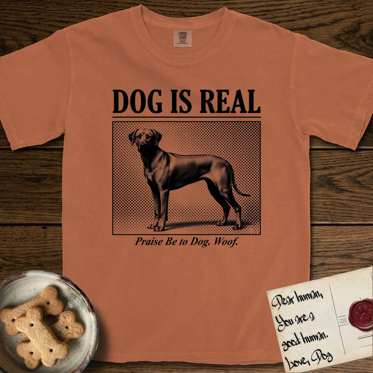 Dog is Real Rhodesian Ridgeback T-Shirt flat lay in Yam, featuring Black halftone newspaper photo of a lean Rhodesian Ridgeback standing in profile with head turned to the camera, under the headline DOG IS REAL, with the caption Praise Be to Dog. Woof..
