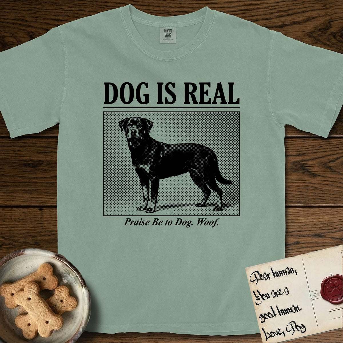 Dog is Real Rottweiler T-Shirt flat lay in Bay, featuring Black halftone newspaper photo of a Rottweiler standing in profile with head turned to the camera, under the headline DOG IS REAL, with the caption Praise Be to Dog. Woof..