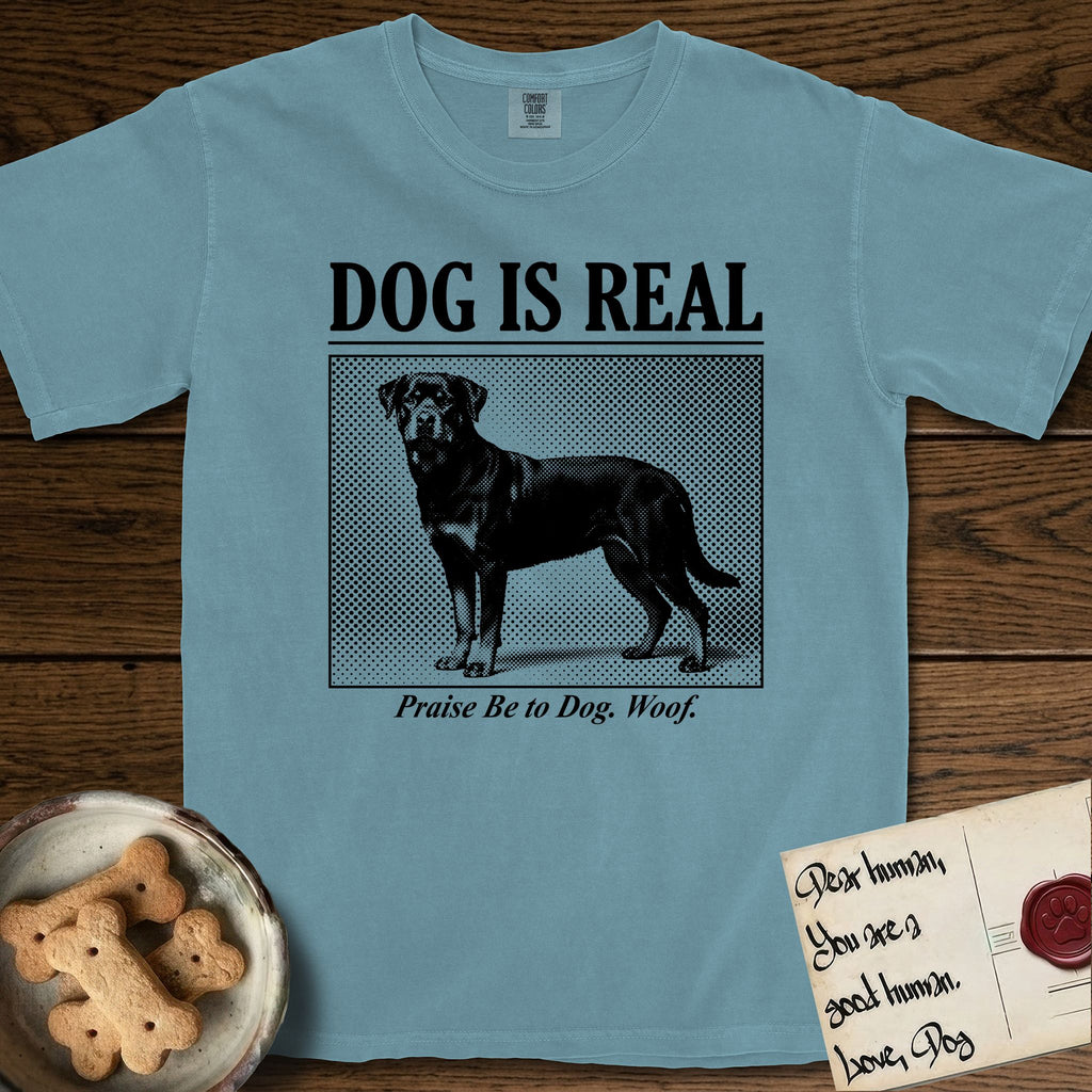 Dog is Real Rottweiler T-Shirt flat lay in Ice Blue, featuring Black halftone newspaper photo of a Rottweiler standing in profile with head turned to the camera, under the headline DOG IS REAL, with the caption Praise Be to Dog. Woof..