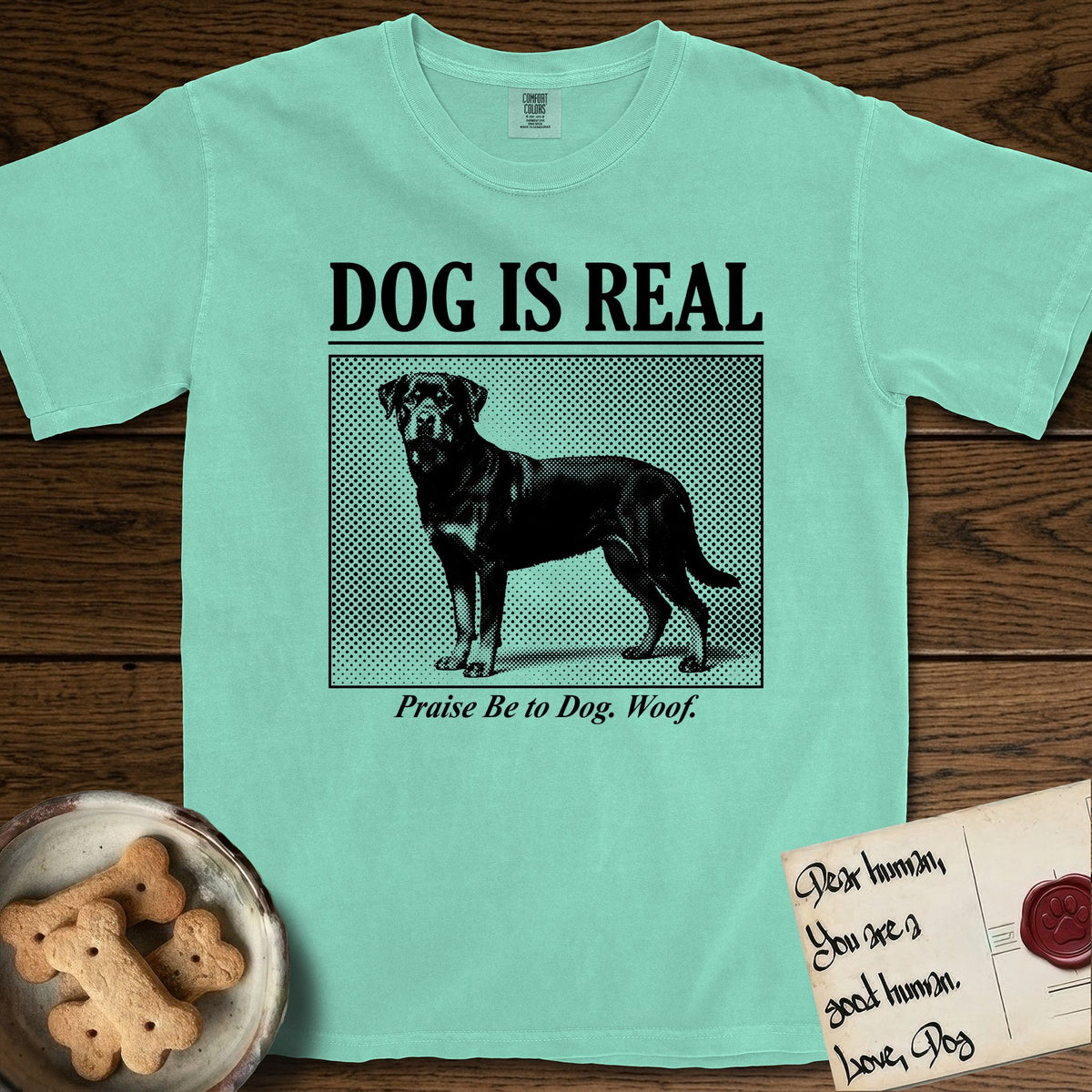 Dog is Real Rottweiler T-Shirt flat lay in Island Reef, featuring Black halftone newspaper photo of a Rottweiler standing in profile with head turned to the camera, under the headline DOG IS REAL, with the caption Praise Be to Dog. Woof..