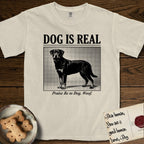Dog is Real Rottweiler T-Shirt flat lay in Ivory, featuring Black halftone newspaper photo of a Rottweiler standing in profile with head turned to the camera, under the headline DOG IS REAL, with the caption Praise Be to Dog. Woof..