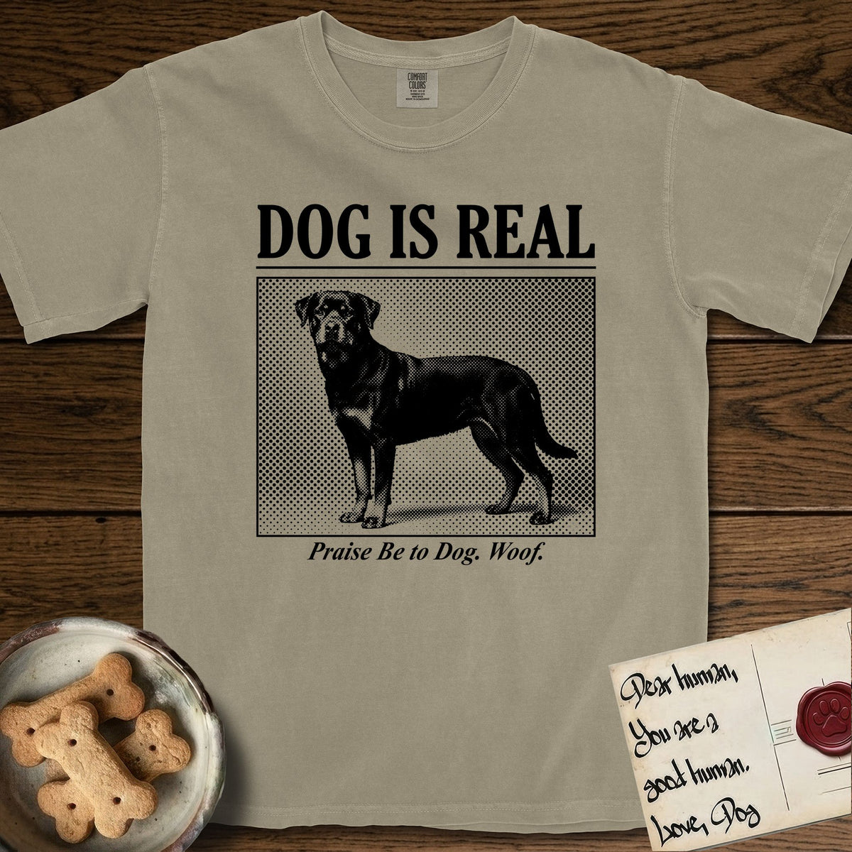 Dog is Real Rottweiler T-Shirt flat lay in Khaki, featuring Black halftone newspaper photo of a Rottweiler standing in profile with head turned to the camera, under the headline DOG IS REAL, with the caption Praise Be to Dog. Woof..