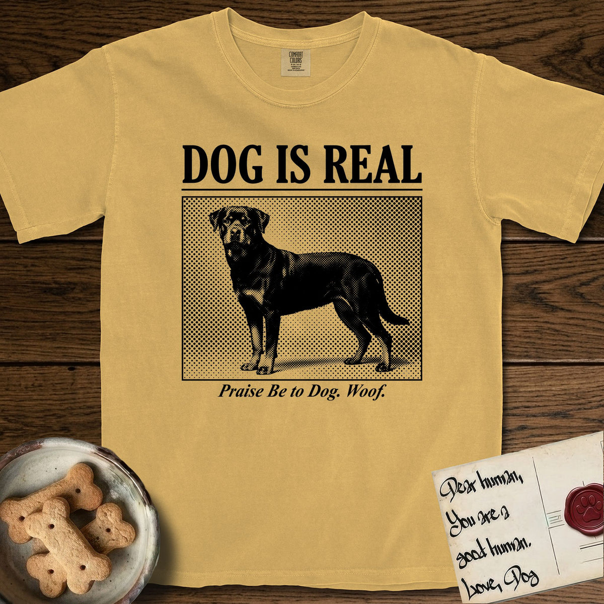 Dog is Real Rottweiler T-Shirt flat lay in Mustard, featuring Black halftone newspaper photo of a Rottweiler standing in profile with head turned to the camera, under the headline DOG IS REAL, with the caption Praise Be to Dog. Woof..