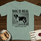 Dog is Real Saint Bernard T-Shirt flat lay in Bay, featuring Black halftone newspaper photo of a Saint Bernard with a white blaze and dark mask standing in profile with head turned to the camera, under the headline DOG IS REAL, with the caption Praise Be to Dog. Woof..