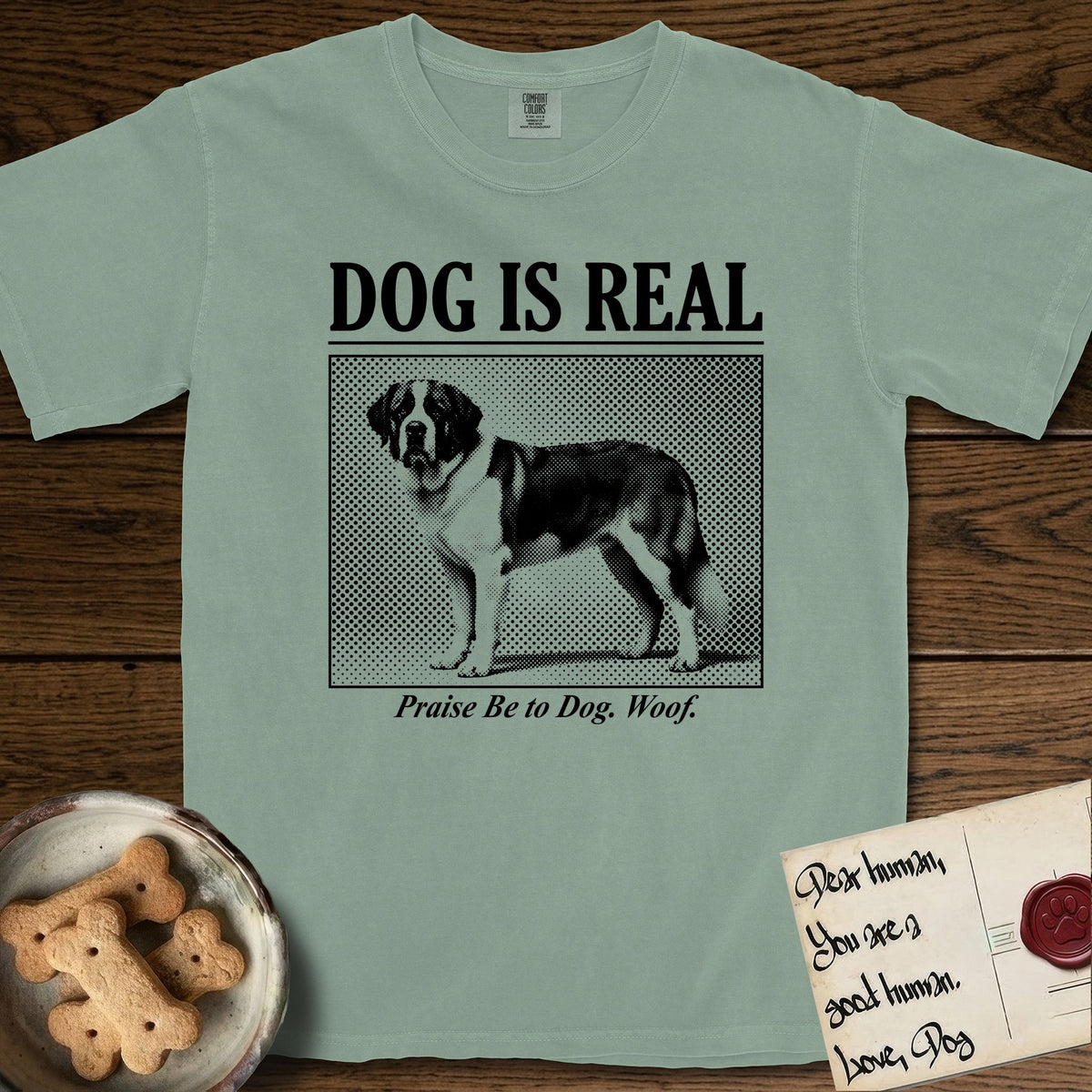 Dog is Real Saint Bernard T-Shirt flat lay in Bay, featuring Black halftone newspaper photo of a Saint Bernard with a white blaze and dark mask standing in profile with head turned to the camera, under the headline DOG IS REAL, with the caption Praise Be to Dog. Woof..