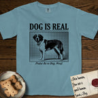 Dog is Real Saint Bernard T-Shirt flat lay in Ice Blue, featuring Black halftone newspaper photo of a Saint Bernard with a white blaze and dark mask standing in profile with head turned to the camera, under the headline DOG IS REAL, with the caption Praise Be to Dog. Woof..