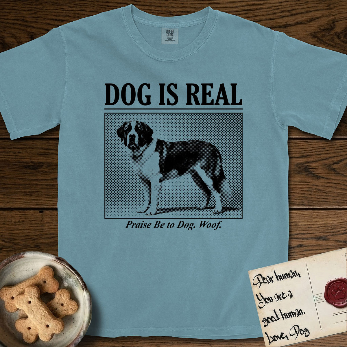 Dog is Real Saint Bernard T-Shirt flat lay in Ice Blue, featuring Black halftone newspaper photo of a Saint Bernard with a white blaze and dark mask standing in profile with head turned to the camera, under the headline DOG IS REAL, with the caption Praise Be to Dog. Woof..