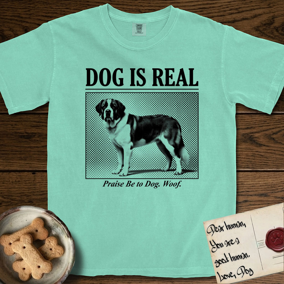Dog is Real Saint Bernard T-Shirt flat lay in Island Reef, featuring Black halftone newspaper photo of a Saint Bernard with a white blaze and dark mask standing in profile with head turned to the camera, under the headline DOG IS REAL, with the caption Praise Be to Dog. Woof..