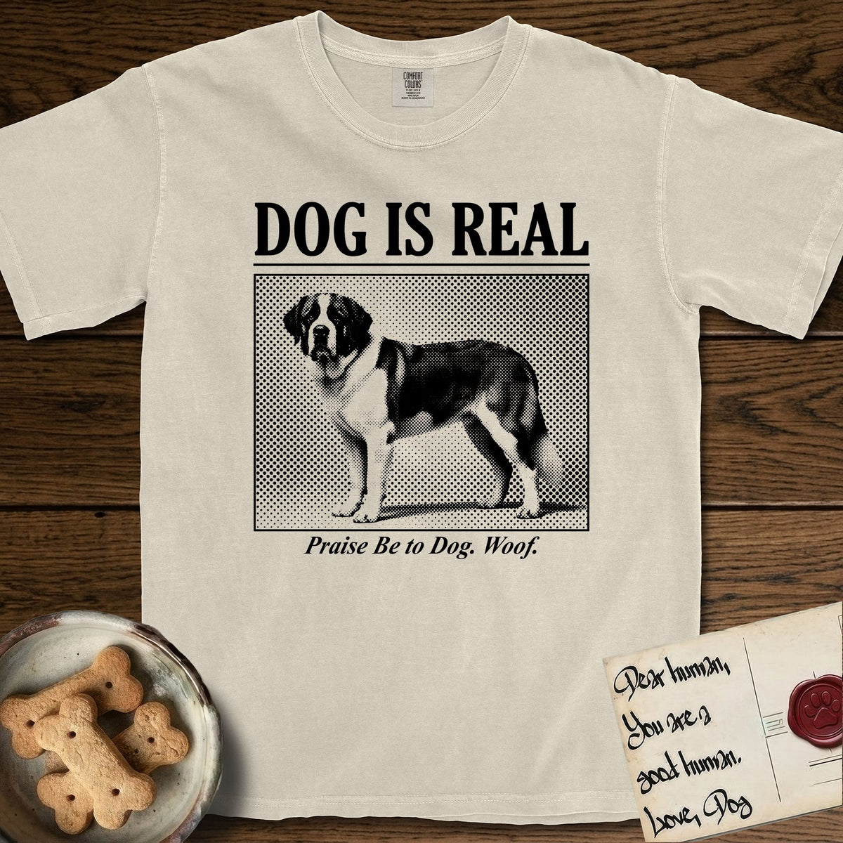 Dog is Real Saint Bernard T-Shirt flat lay in Ivory, featuring Black halftone newspaper photo of a Saint Bernard with a white blaze and dark mask standing in profile with head turned to the camera, under the headline DOG IS REAL, with the caption Praise Be to Dog. Woof..