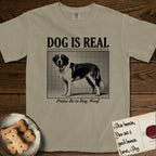 Dog is Real Saint Bernard T-Shirt flat lay in Khaki, featuring Black halftone newspaper photo of a Saint Bernard with a white blaze and dark mask standing in profile with head turned to the camera, under the headline DOG IS REAL, with the caption Praise Be to Dog. Woof..
