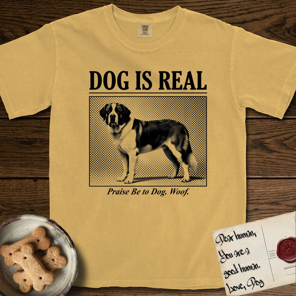 Dog is Real Saint Bernard T-Shirt flat lay in Mustard, featuring Black halftone newspaper photo of a Saint Bernard with a white blaze and dark mask standing in profile with head turned to the camera, under the headline DOG IS REAL, with the caption Praise Be to Dog. Woof..