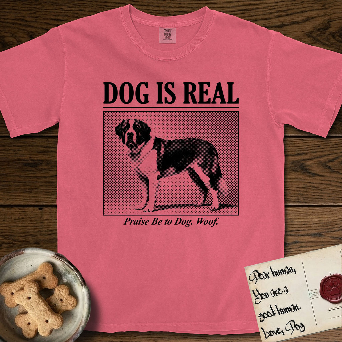 Dog is Real Saint Bernard T-Shirt flat lay in Watermelon, featuring Black halftone newspaper photo of a Saint Bernard with a white blaze and dark mask standing in profile with head turned to the camera, under the headline DOG IS REAL, with the caption Praise Be to Dog. Woof..