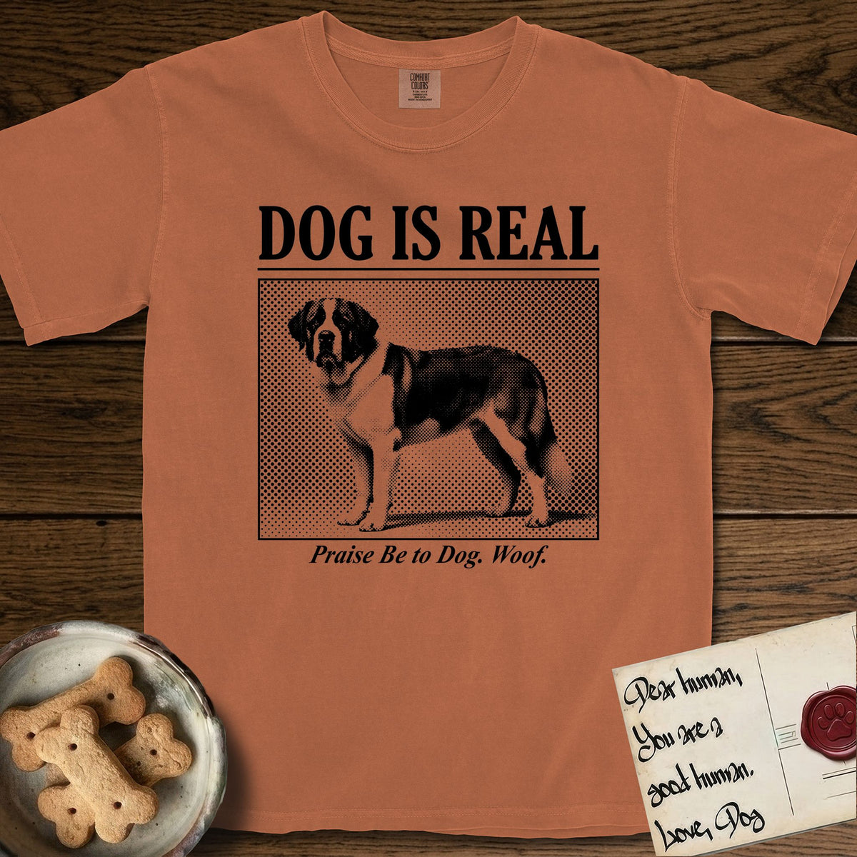 Dog is Real Saint Bernard T-Shirt flat lay in Yam, featuring Black halftone newspaper photo of a Saint Bernard with a white blaze and dark mask standing in profile with head turned to the camera, under the headline DOG IS REAL, with the caption Praise Be to Dog. Woof..