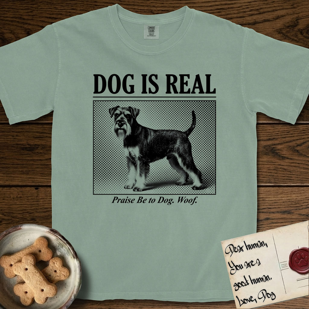 Dog is Real Schnauzer T-Shirt flat lay in Bay, featuring Black halftone newspaper photo of a bearded Schnauzer with an upright tail standing in profile with head turned to the camera, under the headline DOG IS REAL, with the caption Praise Be to Dog. Woof..