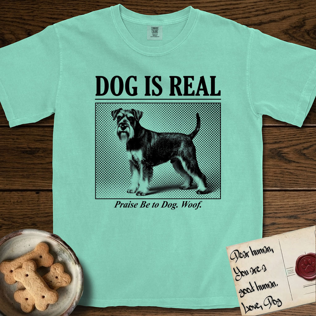 Dog is Real Schnauzer T-Shirt flat lay in Island Reef, featuring Black halftone newspaper photo of a bearded Schnauzer with an upright tail standing in profile with head turned to the camera, under the headline DOG IS REAL, with the caption Praise Be to Dog. Woof..