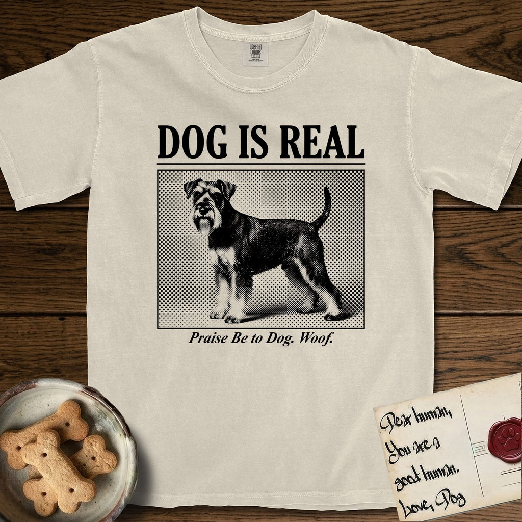 Dog is Real Schnauzer T-Shirt flat lay in Ivory, featuring Black halftone newspaper photo of a bearded Schnauzer with an upright tail standing in profile with head turned to the camera, under the headline DOG IS REAL, with the caption Praise Be to Dog. Woof..
