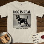 Dog is Real Schnauzer T-Shirt flat lay in Ivory, featuring Black halftone newspaper photo of a bearded Schnauzer with an upright tail standing in profile with head turned to the camera, under the headline DOG IS REAL, with the caption Praise Be to Dog. Woof..