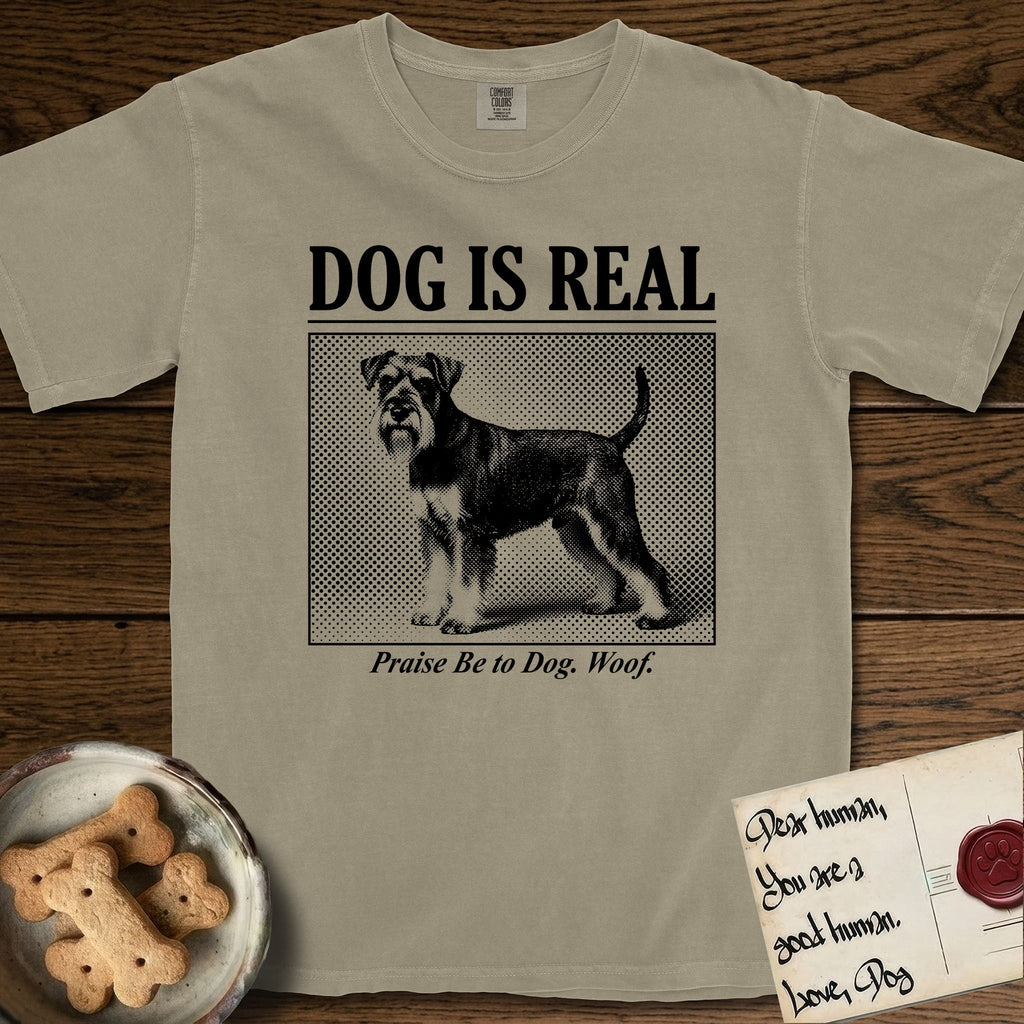 Dog is Real Schnauzer T-Shirt flat lay in Khaki, featuring Black halftone newspaper photo of a bearded Schnauzer with an upright tail standing in profile with head turned to the camera, under the headline DOG IS REAL, with the caption Praise Be to Dog. Woof..