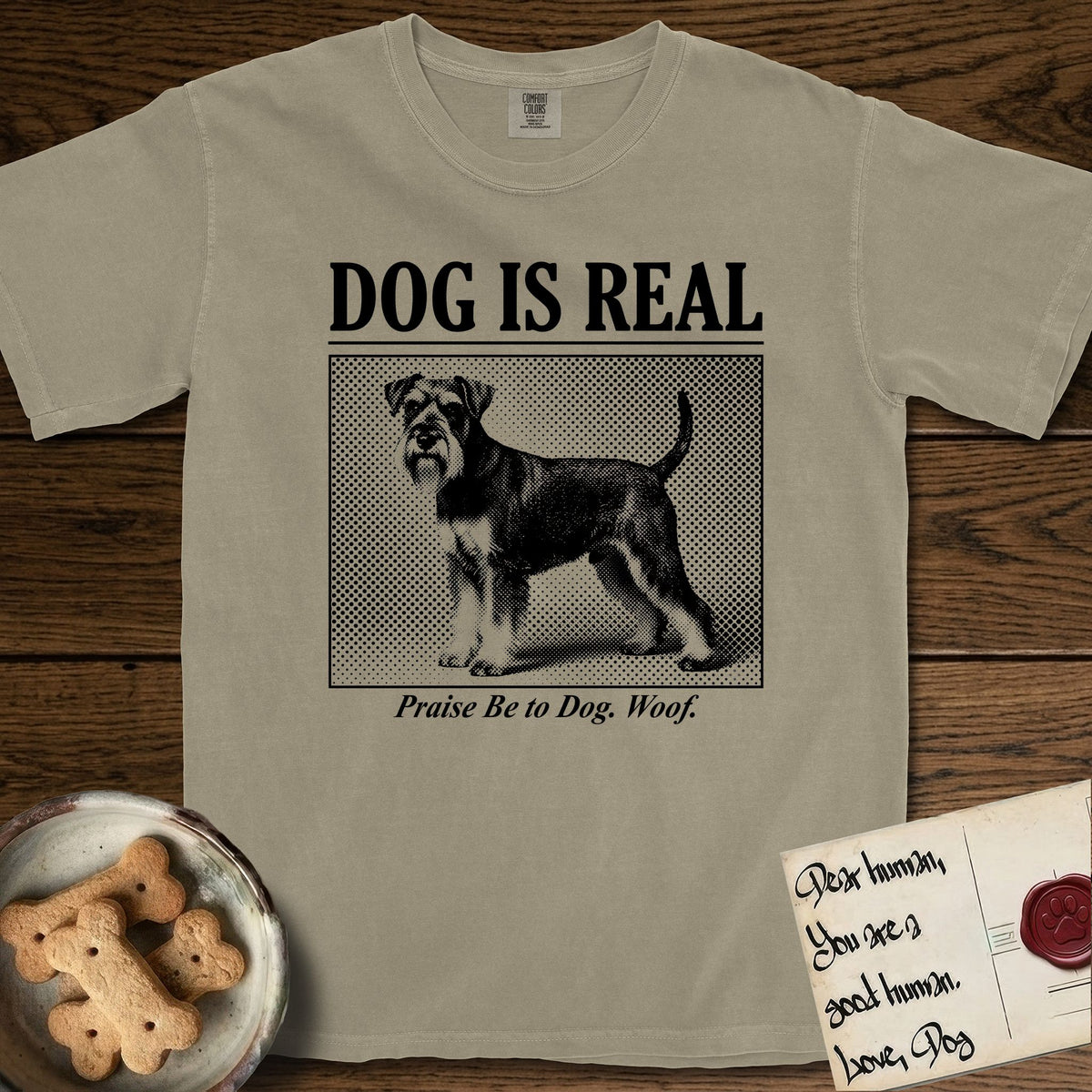 Dog is Real Schnauzer T-Shirt flat lay in Khaki, featuring Black halftone newspaper photo of a bearded Schnauzer with an upright tail standing in profile with head turned to the camera, under the headline DOG IS REAL, with the caption Praise Be to Dog. Woof..