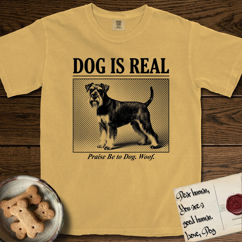 Dog is Real Schnauzer T-Shirt flat lay in Mustard, featuring Black halftone newspaper photo of a bearded Schnauzer with an upright tail standing in profile with head turned to the camera, under the headline DOG IS REAL, with the caption Praise Be to Dog. Woof..
