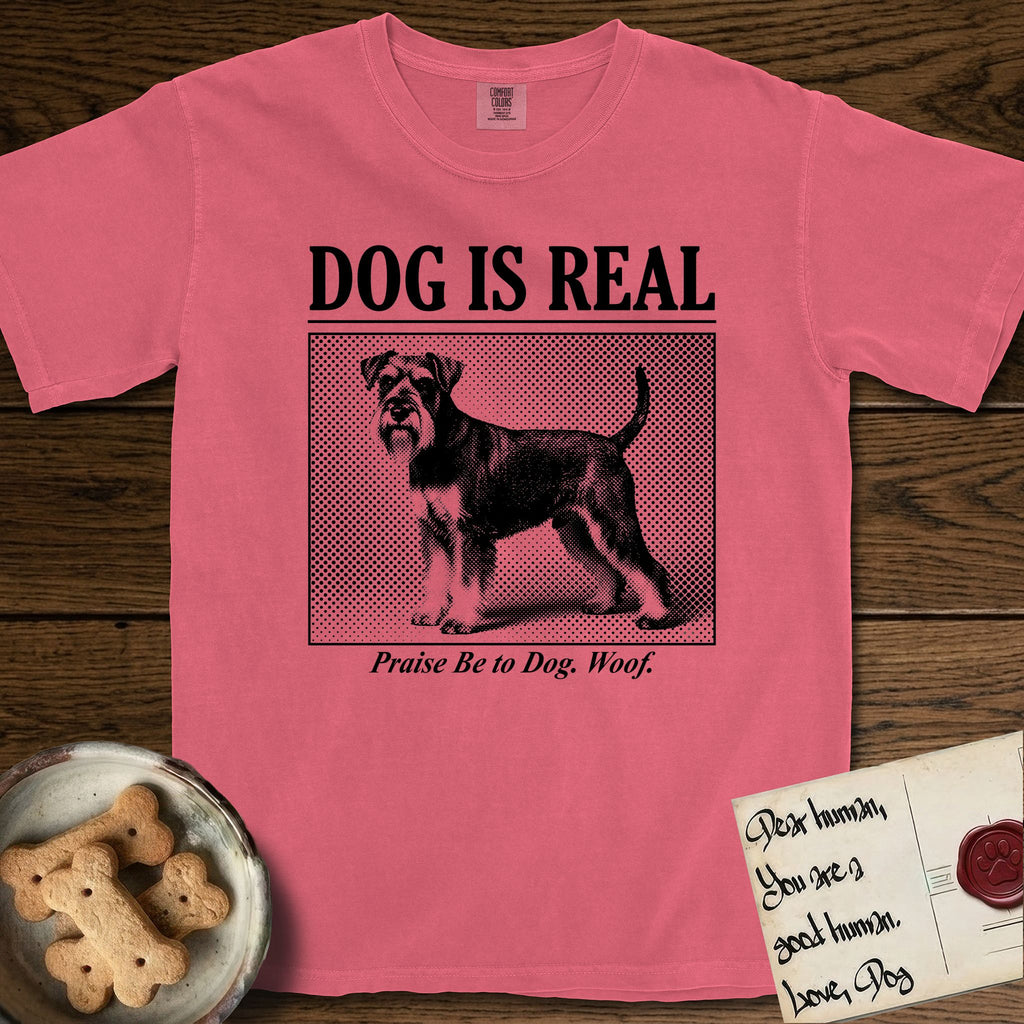 Dog is Real Schnauzer T-Shirt flat lay in Watermelon, featuring Black halftone newspaper photo of a bearded Schnauzer with an upright tail standing in profile with head turned to the camera, under the headline DOG IS REAL, with the caption Praise Be to Dog. Woof..