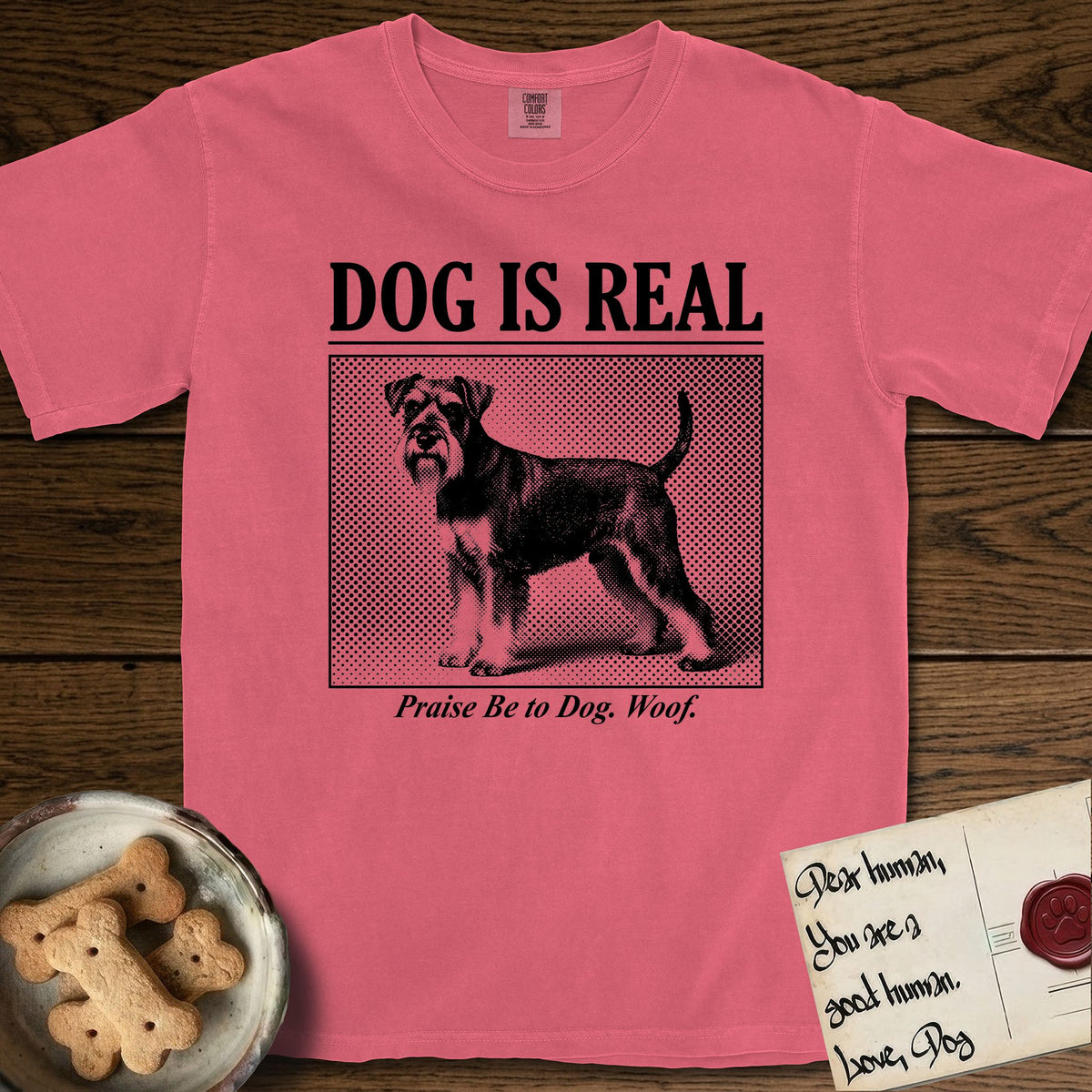 Dog is Real Schnauzer T-Shirt flat lay in Watermelon, featuring Black halftone newspaper photo of a bearded Schnauzer with an upright tail standing in profile with head turned to the camera, under the headline DOG IS REAL, with the caption Praise Be to Dog. Woof..