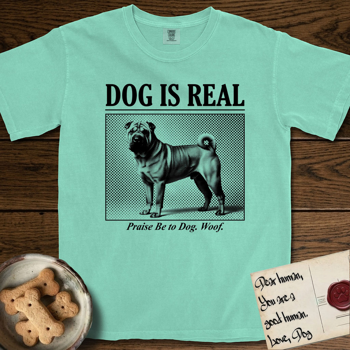 Dog is Real Shar-Pei T-Shirt flat lay in Island Reef, featuring Black halftone newspaper photo of a wrinkled Shar-Pei with a curled tail standing in profile with head turned to the camera, under the headline DOG IS REAL, with the caption Praise Be to Dog. Woof..