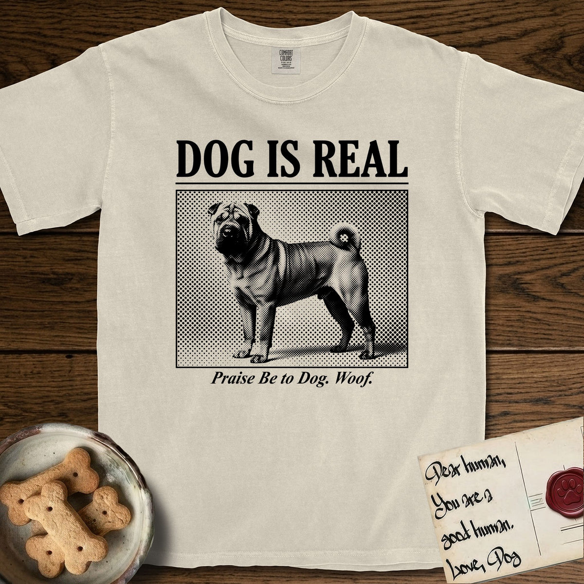 Dog is Real Shar-Pei T-Shirt flat lay in Ivory, featuring Black halftone newspaper photo of a wrinkled Shar-Pei with a curled tail standing in profile with head turned to the camera, under the headline DOG IS REAL, with the caption Praise Be to Dog. Woof..