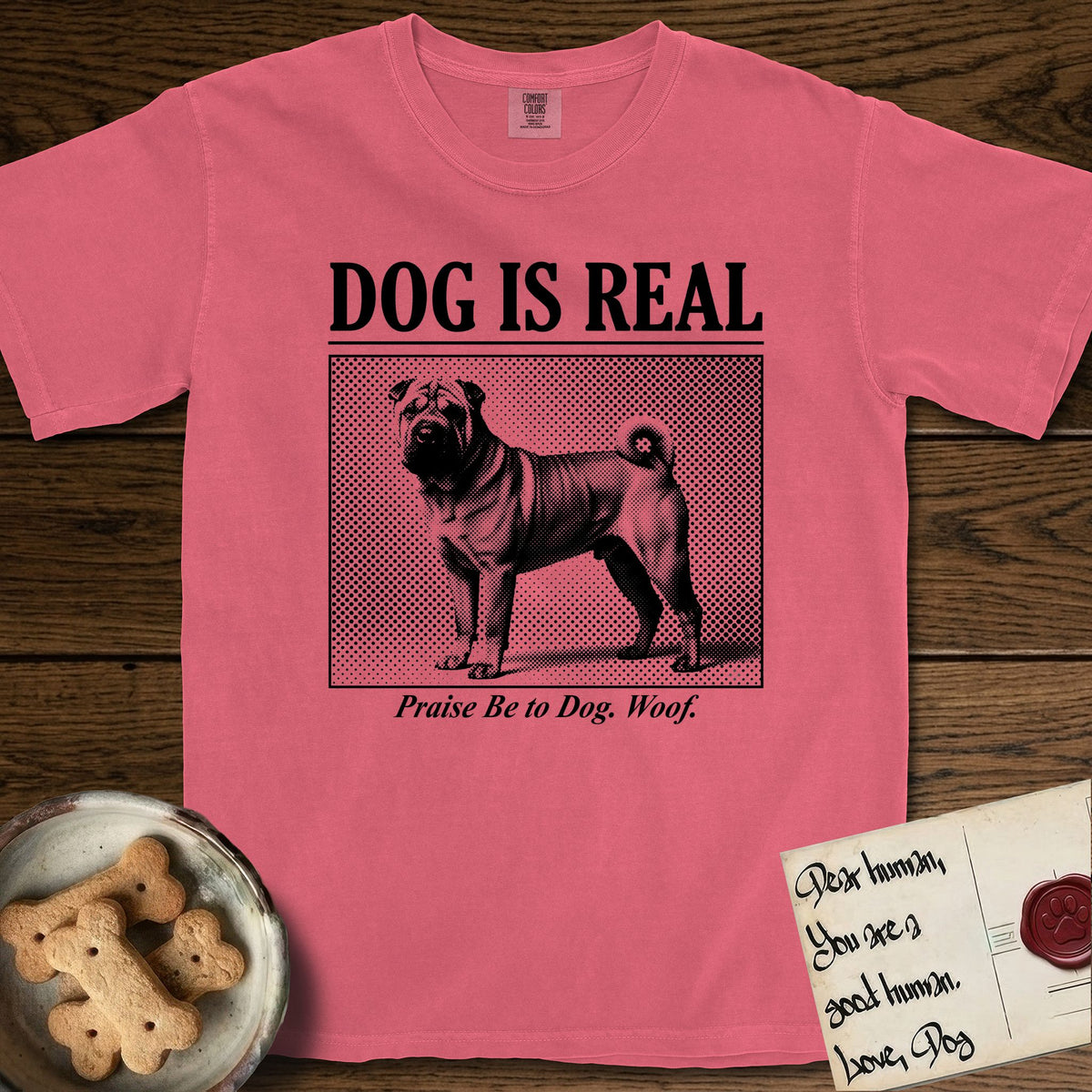 Dog is Real Shar-Pei T-Shirt flat lay in Watermelon, featuring Black halftone newspaper photo of a wrinkled Shar-Pei with a curled tail standing in profile with head turned to the camera, under the headline DOG IS REAL, with the caption Praise Be to Dog. Woof..