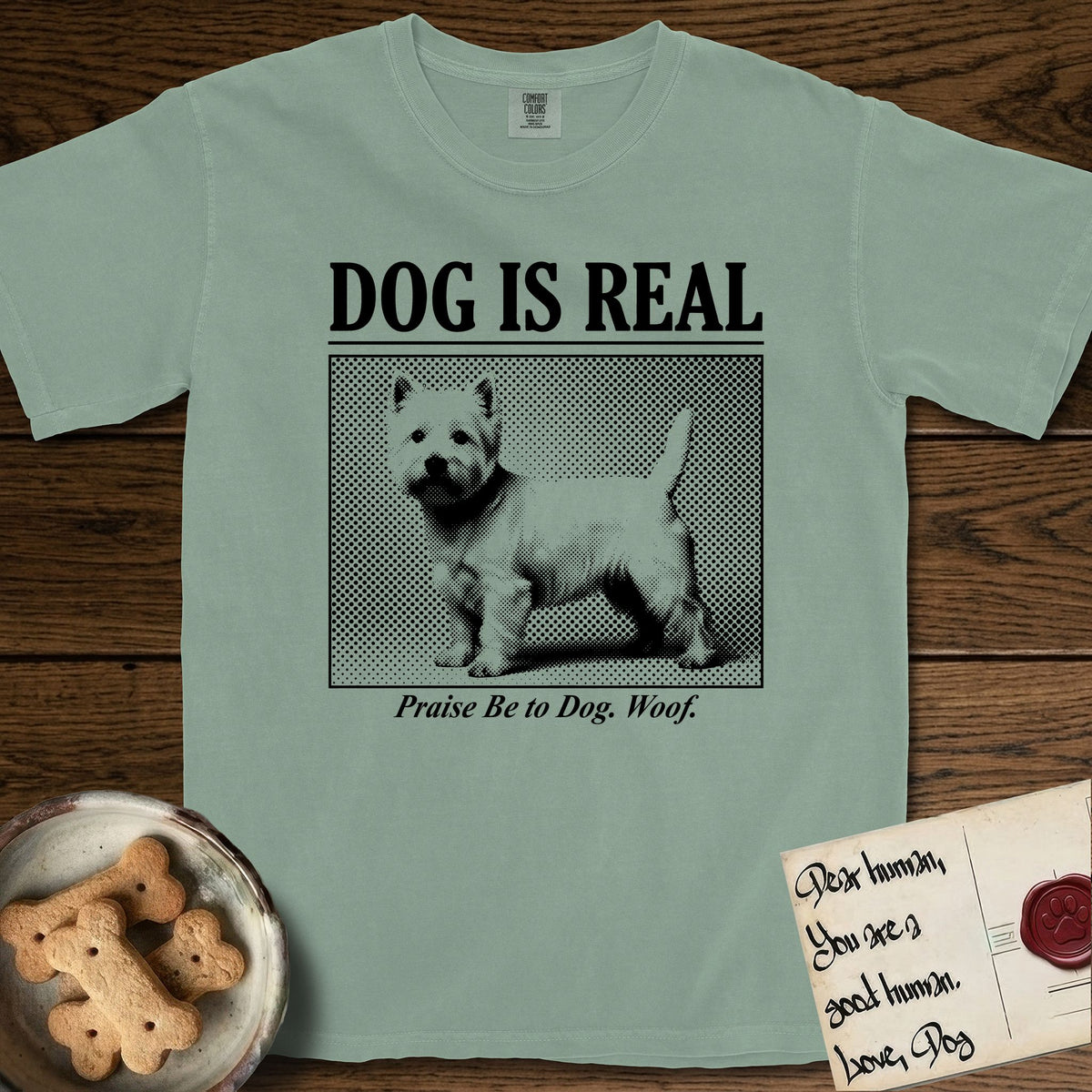 Dog is Real Westie T-Shirt flat lay in Bay, featuring Black halftone newspaper photo of a white Westie with upright ears and tail standing in profile with head turned to the camera, under the headline DOG IS REAL, with the caption Praise Be to Dog. Woof..