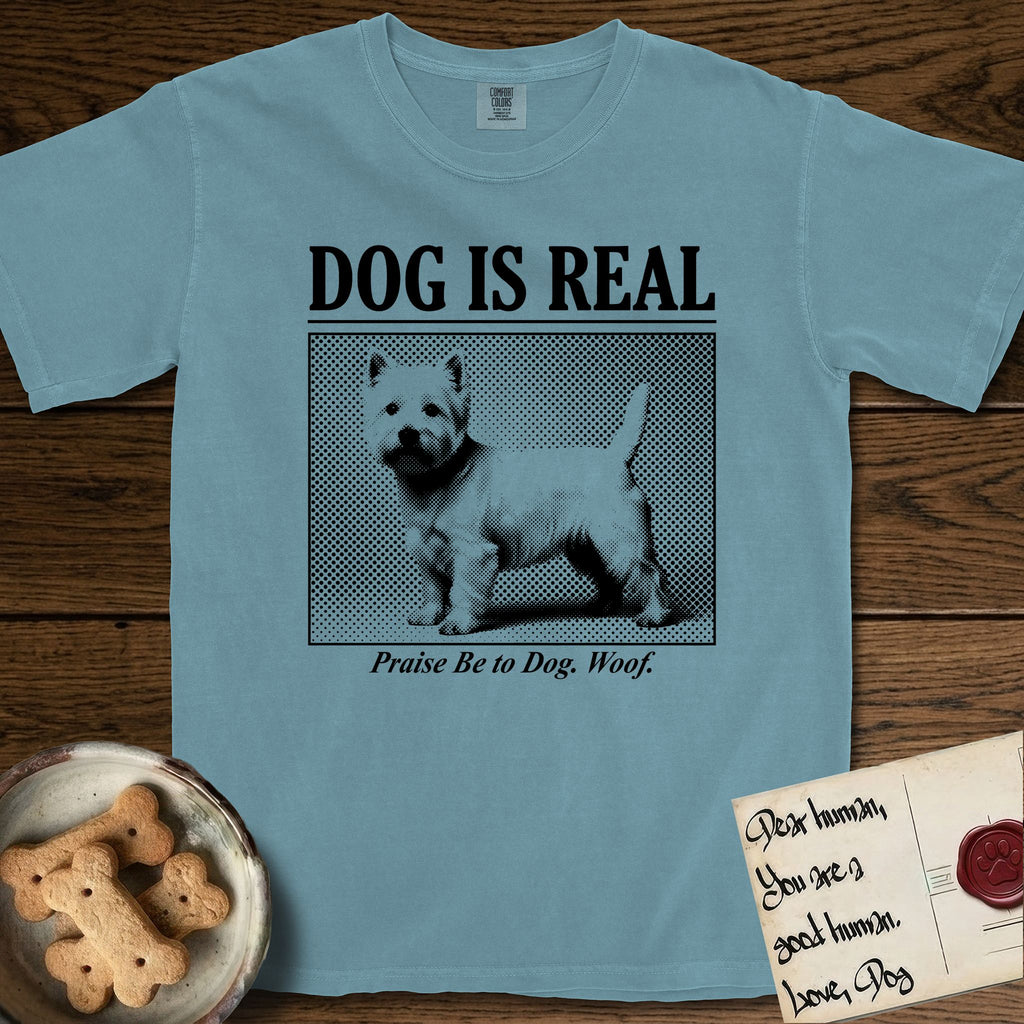 Dog is Real Westie T-Shirt flat lay in Ice Blue, featuring Black halftone newspaper photo of a white Westie with upright ears and tail standing in profile with head turned to the camera, under the headline DOG IS REAL, with the caption Praise Be to Dog. Woof..
