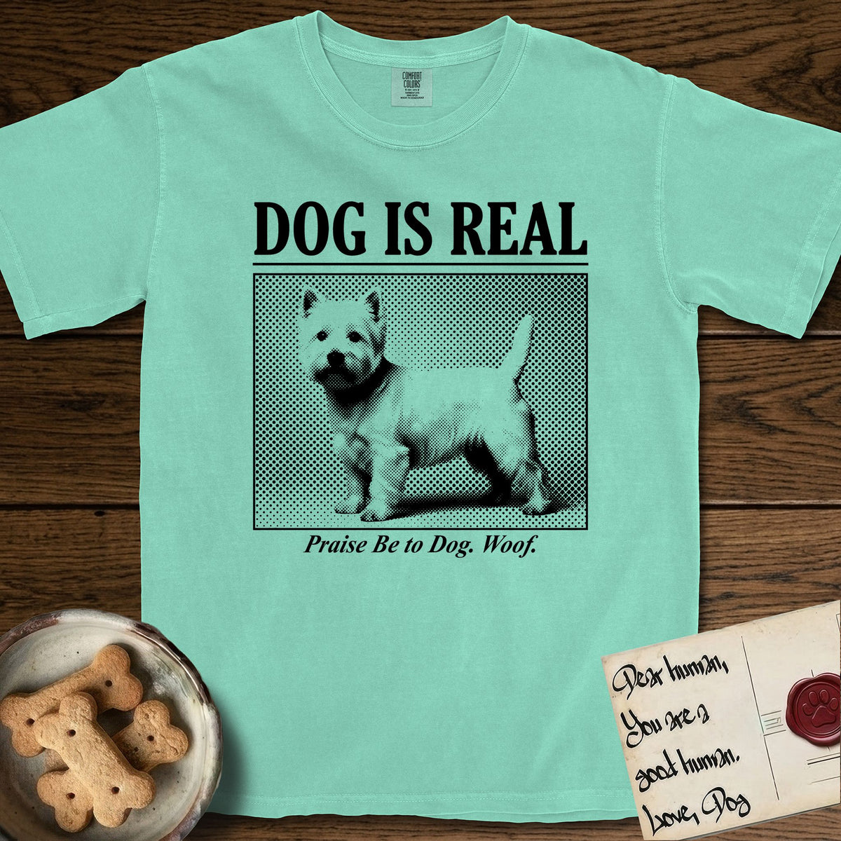Dog is Real Westie T-Shirt flat lay in Island Reef, featuring Black halftone newspaper photo of a white Westie with upright ears and tail standing in profile with head turned to the camera, under the headline DOG IS REAL, with the caption Praise Be to Dog. Woof..