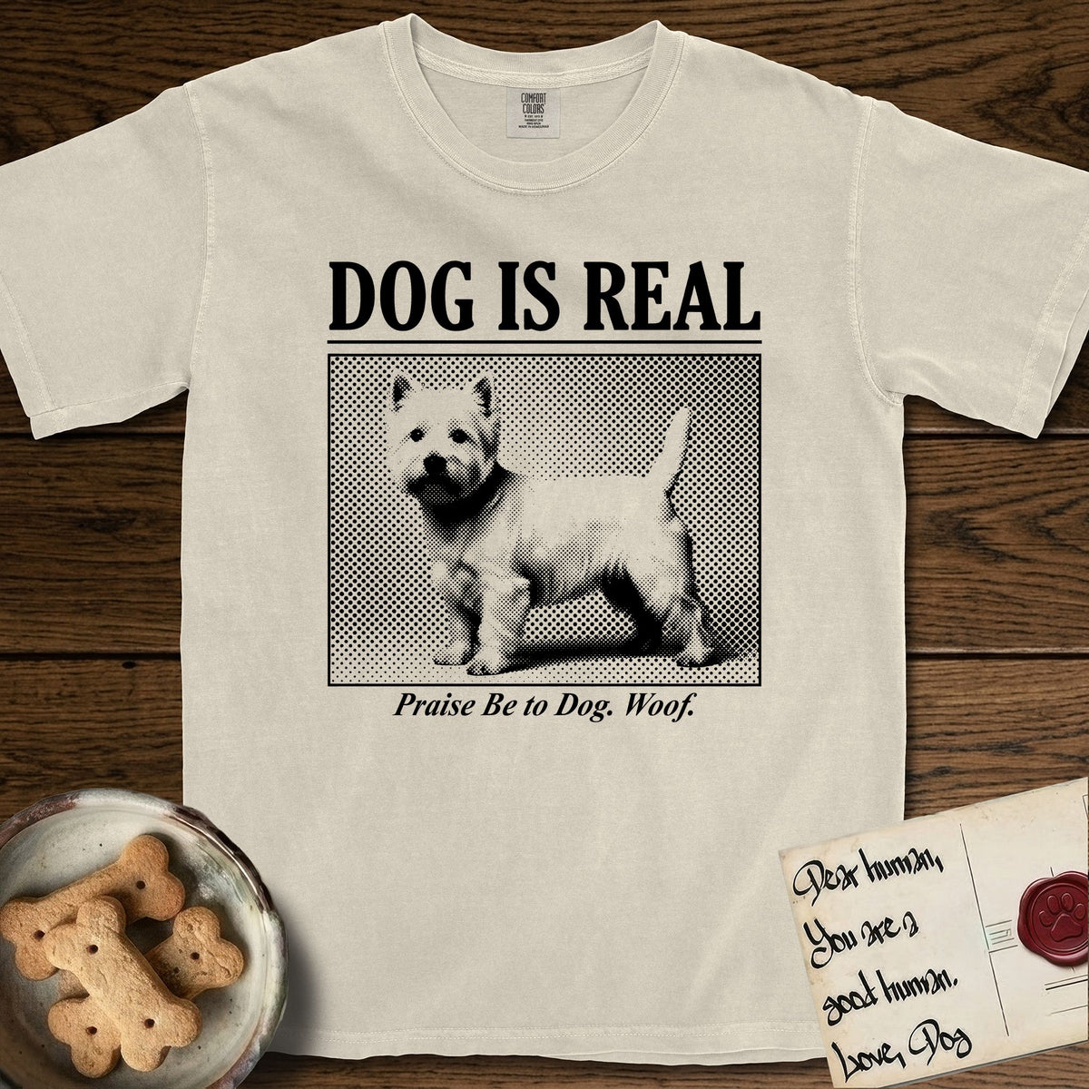 Dog is Real Westie T-Shirt flat lay in Ivory, featuring Black halftone newspaper photo of a white Westie with upright ears and tail standing in profile with head turned to the camera, under the headline DOG IS REAL, with the caption Praise Be to Dog. Woof..
