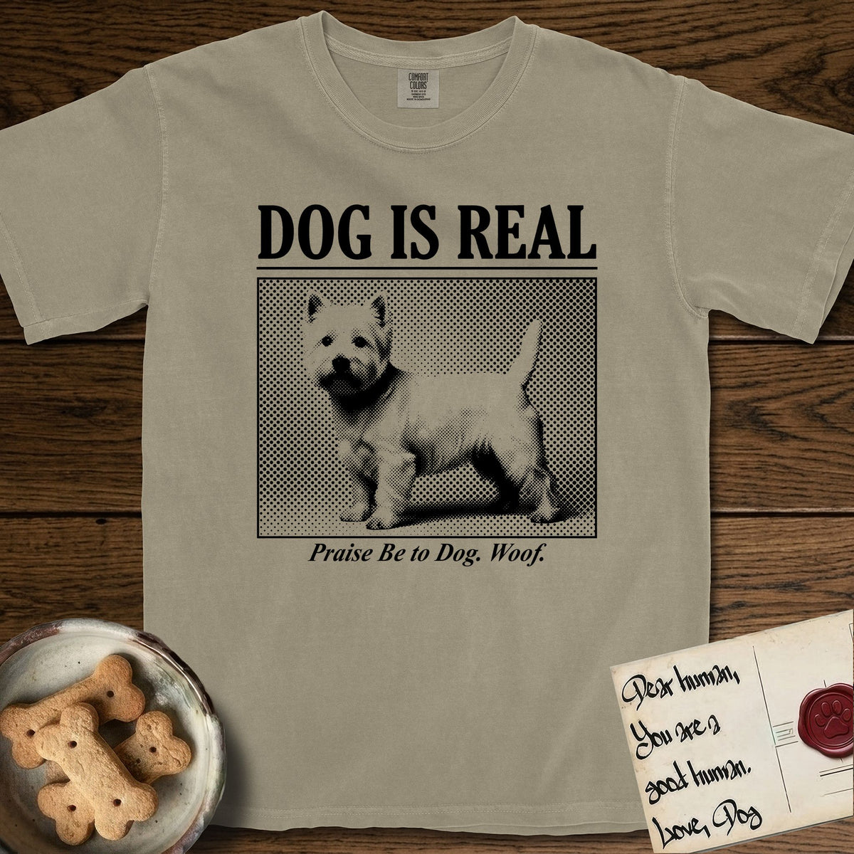 Dog is Real Westie T-Shirt flat lay in Khaki, featuring Black halftone newspaper photo of a white Westie with upright ears and tail standing in profile with head turned to the camera, under the headline DOG IS REAL, with the caption Praise Be to Dog. Woof..