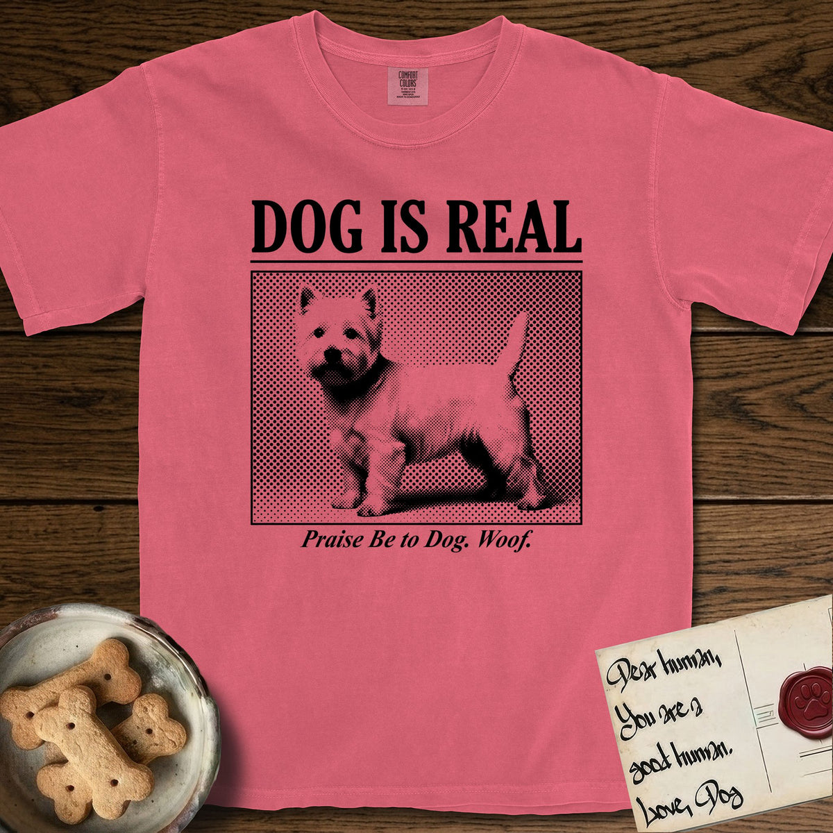 Dog is Real Westie T-Shirt flat lay in Watermelon, featuring Black halftone newspaper photo of a white Westie with upright ears and tail standing in profile with head turned to the camera, under the headline DOG IS REAL, with the caption Praise Be to Dog. Woof..