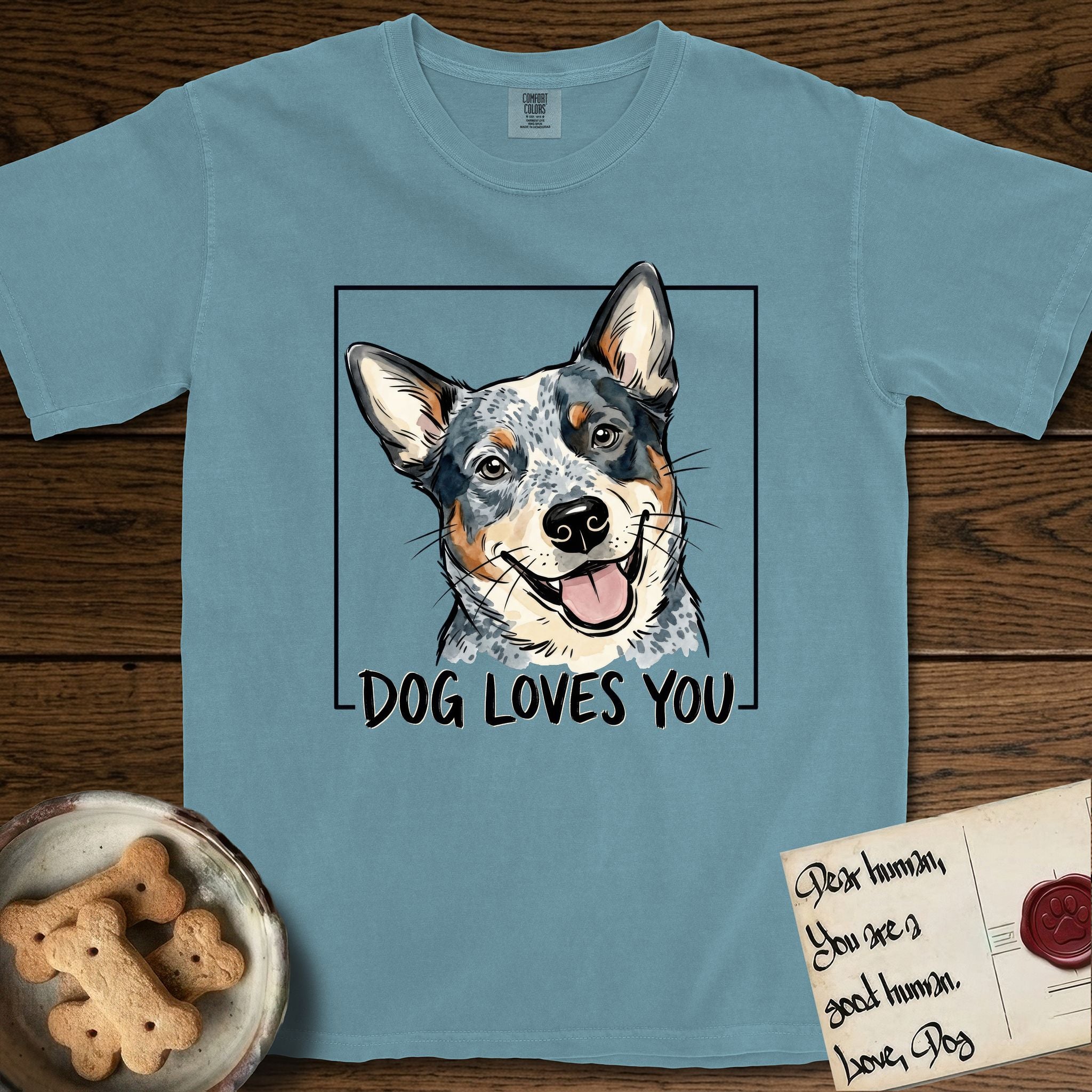 Dog Loves You Australian Cattle Dog T-Shirt flat lay in Ice Blue, featuring Ink and watercolor portrait of a smiling blue Australian Cattle Dog with a speckled coat, tan eyebrows and upright ears, head tilted inside a black frame above hand-lettered text reading DOG LOVES YOU.