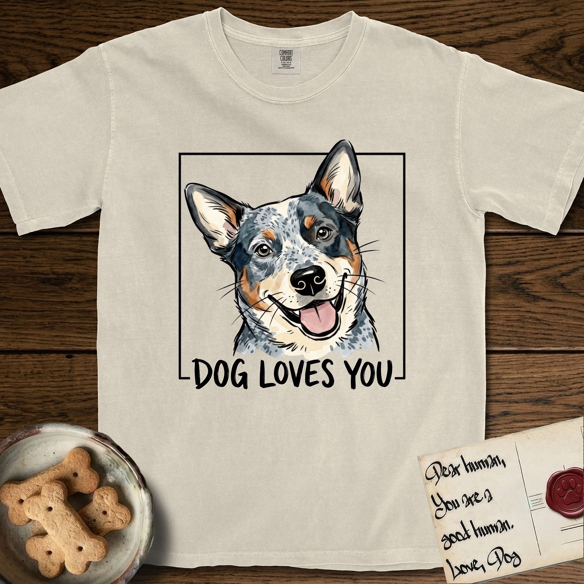 Dog Loves You Australian Cattle Dog T-Shirt flat lay in Ivory, featuring Ink and watercolor portrait of a smiling blue Australian Cattle Dog with a speckled coat, tan eyebrows and upright ears, head tilted inside a black frame above hand-lettered text reading DOG LOVES YOU.