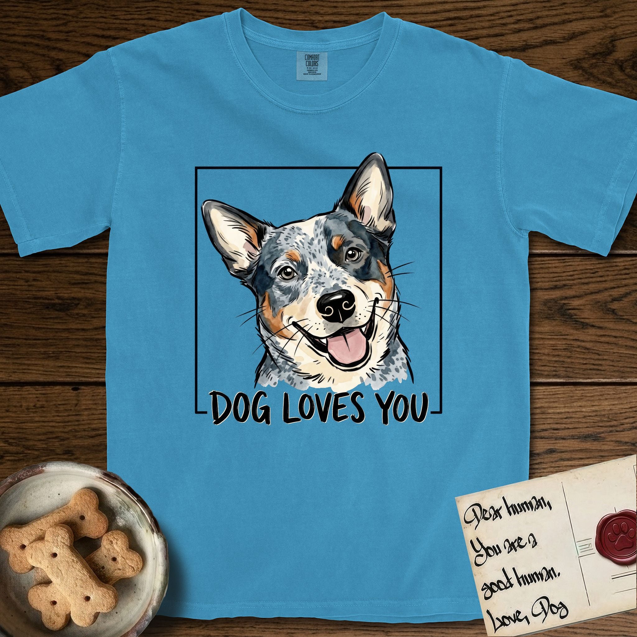 Dog Loves You Australian Cattle Dog T-Shirt flat lay in Royal Caribe, featuring Ink and watercolor portrait of a smiling blue Australian Cattle Dog with a speckled coat, tan eyebrows and upright ears, head tilted inside a black frame above hand-lettered text reading DOG LOVES YOU.