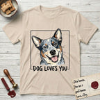 Dog Loves You Australian Cattle Dog T-Shirt flat lay in Sand, featuring Ink and watercolor portrait of a smiling blue Australian Cattle Dog with a speckled coat, tan eyebrows and upright ears, head tilted inside a black frame above hand-lettered text reading DOG LOVES YOU.