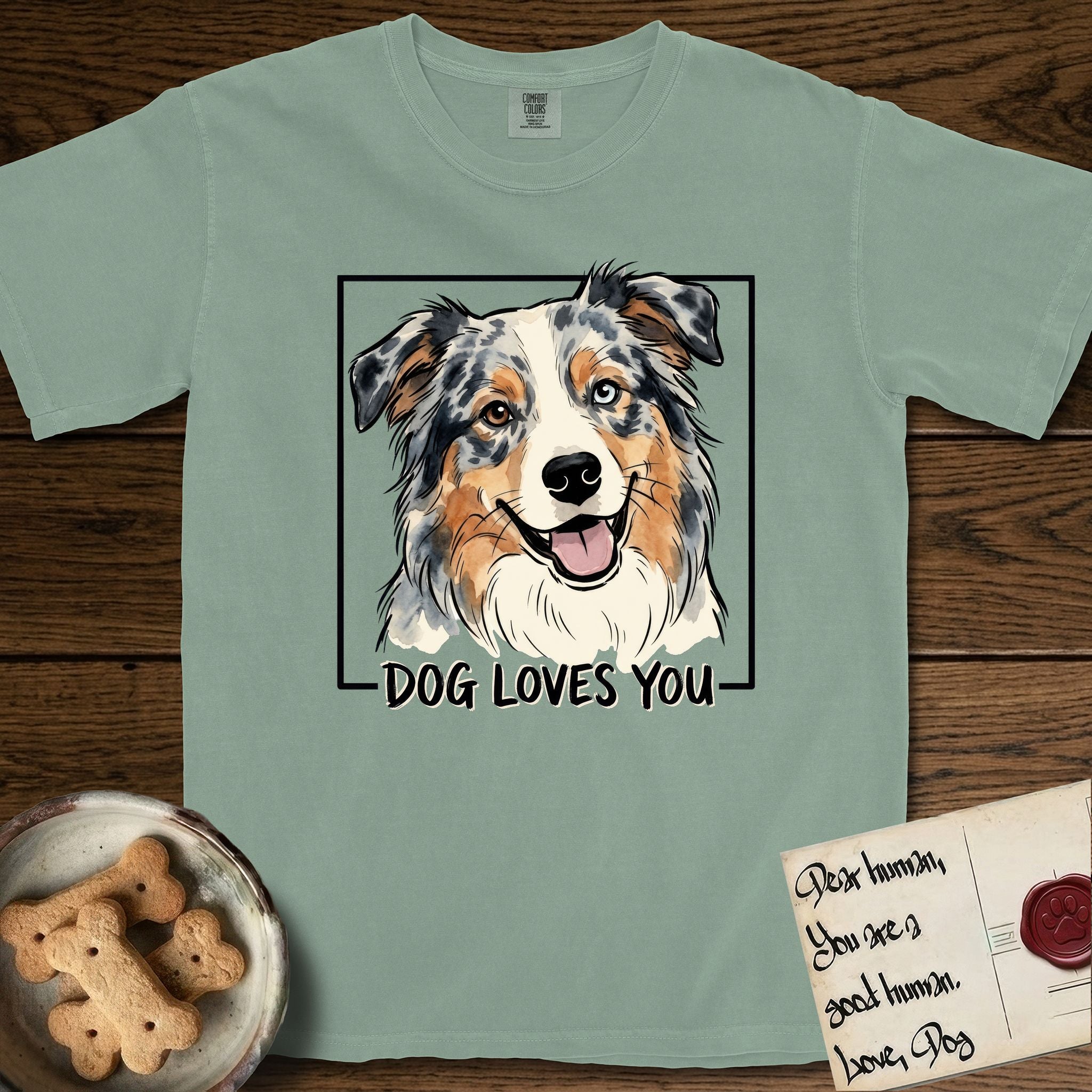 Dog Loves You Australian Shepherd T-Shirt flat lay in Bay, featuring Ink and watercolor portrait of a smiling blue merle Australian Shepherd with one brown eye and one blue eye and a cream blaze, head tilted inside a black frame above hand-lettered text reading DOG LOVES YOU.