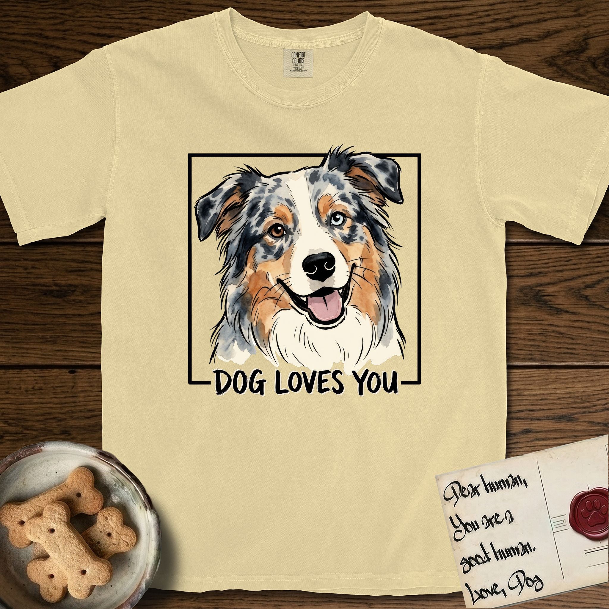 Dog Loves You Australian Shepherd T-Shirt flat lay in Butter, featuring Ink and watercolor portrait of a smiling blue merle Australian Shepherd with one brown eye and one blue eye and a cream blaze, head tilted inside a black frame above hand-lettered text reading DOG LOVES YOU.