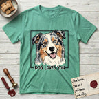 Dog Loves You Australian Shepherd T-Shirt flat lay in Heather Irish Green, featuring Ink and watercolor portrait of a smiling blue merle Australian Shepherd with one brown eye and one blue eye and a cream blaze, head tilted inside a black frame above hand-lettered text reading DOG LOVES YOU.